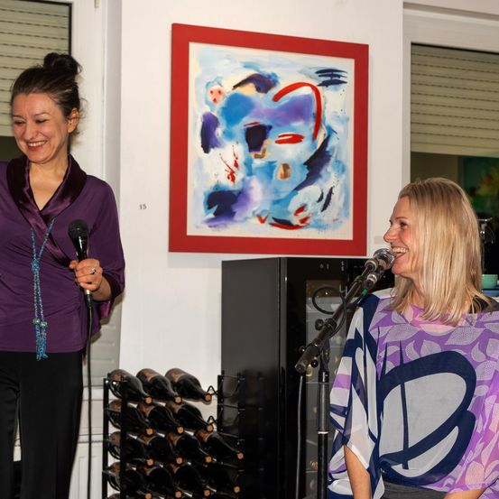 Zwei Frauen stehen, eine hält ein Mikrofon und lächelt, die andere singt in ein Mikrofon. Ein abstraktes Gemälde an der Wand, darunter ein Weinregal.