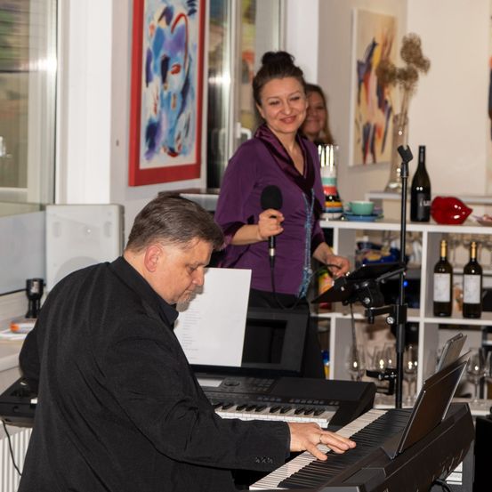 Ein Mann spielt Klavier, während eine Frau mit einem Mikrofon lächelt und hinter ihm steht. Hinter der Frau befindet sich ein Regal mit Flaschen und anderen Gegenständen.