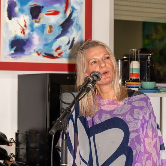 Eine Frau mit blondem Haar spricht in ein Mikrofon mit einem purpurfarbenen und weißen gemusterten Umhang. Dahinter befindet sich eine Wand mit einem abstrakten Gemälde und einem Regal mit Kaffeemaschine und Tassen.