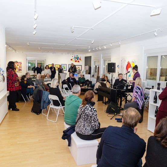 Eine Gruppe von Menschen ist in einem Innenraum versammelt, wahrscheinlich in einer Kunstgalerie. Einige sitzen auf Stühlen, während andere stehen und reden. Es gibt Gemälde an der Wand und eine Person steht vor einem Klavier.