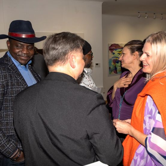 Vier Menschen in einem Raum, zwei Männer und zwei Frauen, wobei ein Mann einen Hut trägt und die anderen lächeln, scheinen sich zu unterhalten.