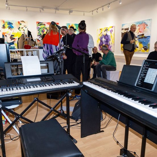 Eine Gruppe von Menschen befindet sich in einer Kunstgalerie. Sie stehen herum und betrachten abstrakte Gemälde. Einige sitzen mit Weingläsern und ein Klavier ist mit Musikblättern aufgestellt.