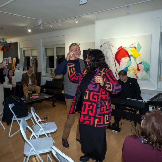Ein Raum mit mehreren Personen, eine Frau in einem gemusterten Kleid tanzt mit einem Mann in einem Pullover, andere sitzen um einen Tisch herum und ein Klavier steht auf dem Boden.