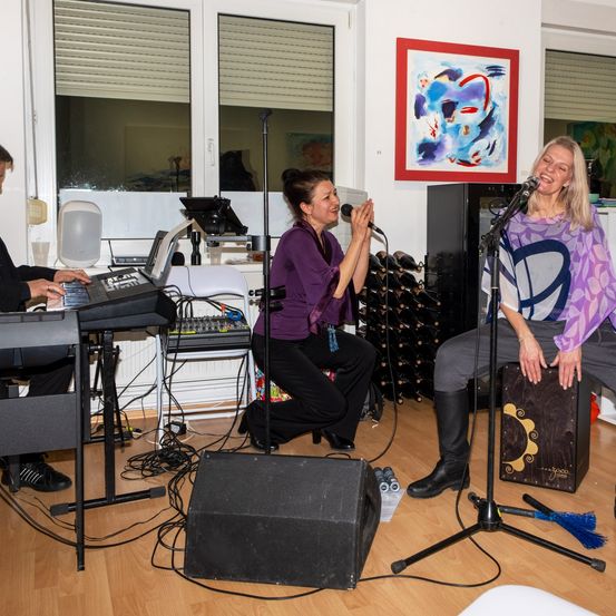 Drei Musiker spielen drinnen; eine Frau singt in ein Mikrofon, eine andere spielt eine Tastatur und ein Mann auf der linken Seite spielt Klavier. Dahinter steht ein Lautsprecher, und an der Wand befindet sich ein abstraktes Gemälde.