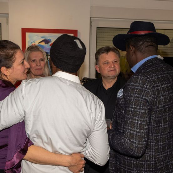 Fünf Menschen sind in einem Raum versammelt und unterhalten sich. Ein Mann in einem weißen Hemd und einer Mütze schaut einen anderen Mann in einem karierten Mantel und Hut an. Eine Frau in Lila und eine andere mit blonden Haaren lauschen. Ein Mann in einem schwarzen Hemd hält ein Glas.