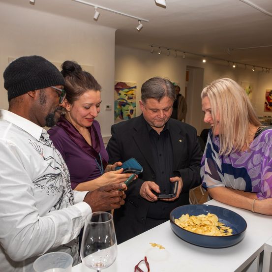 Vier Menschen sind bei einer Veranstaltung, lächeln und schauen auf ihre Handys. Ein Tisch mit Snacks, Gläsern und Tellern ist in der Nähe. Der Raum hat weiße Wände, Deckenleuchten und Kunst an der Wand.