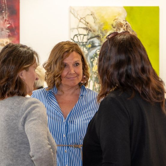 Drei Frauen stehen vor einer Kunstgalerie und unterhalten sich. Die Frau in der Mitte trägt ein blaues Hemd.