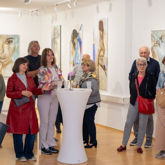 Mehrere Personen stehen in einer modernen Kunstgalerie und bewundern die Gemälde. Ein Tisch mit Gläsern steht in der Mitte, und die Besucher unterhalten sich.