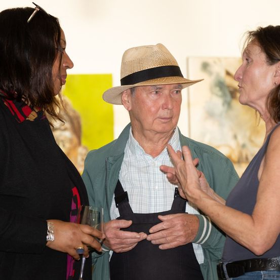 Ein älterer Mann mit Hut und Schürze unterhält sich mit zwei Frauen, eine hält ein Weinglas, in einer Kunstgalerie.