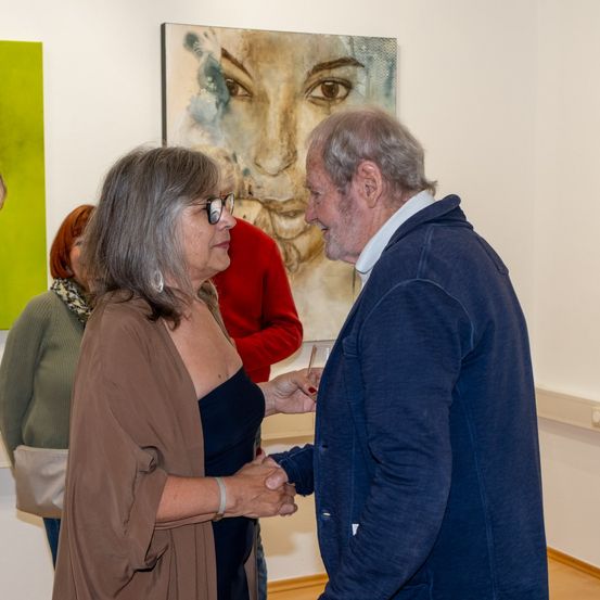 Ein älterer Mann und eine ältere Frau geben sich in einer Kunstgalerie die Hand, hinter ihnen ein großes Porträt. Eine weitere Person steht in der Nähe, und ein grünes Gemälde befindet sich links.