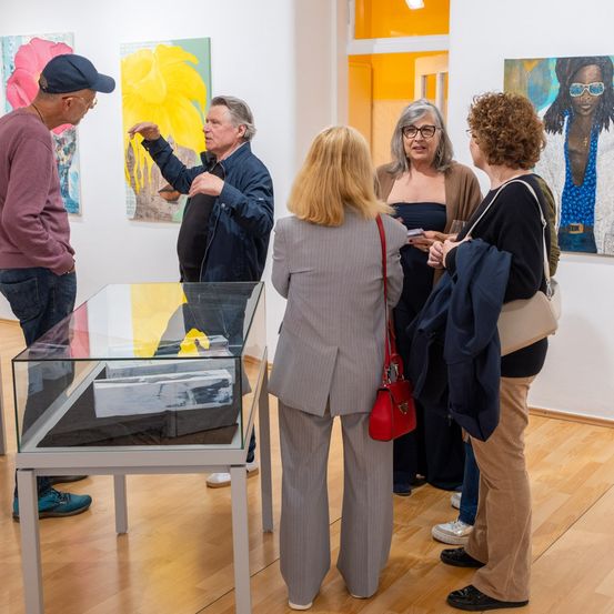 Eine Gruppe von Menschen steht um eine Glasvitrine in einer Galerie und diskutiert über die ausgestellten Kunstwerke. Ein Mann zeigt auf ein Gemälde an der Wand.