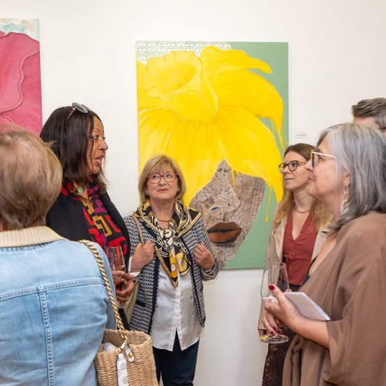 Eine Gruppe von Menschen diskutiert Kunst bei einer Ausstellung, einer hält Wein und eine andere liest. In der Nähe dominiert ein großes Gemälde einer gelben Blume die Wand.