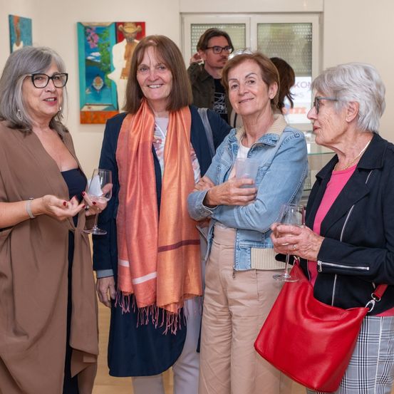 Vier Frauen stehen in einer Kunstgalerie zusammen, lächeln und halten Weingläser in den Händen. Sie tragen lässige Kleidung, eine Frau trägt eine Schürze. Kunstwerke schmücken die weißen Wände.