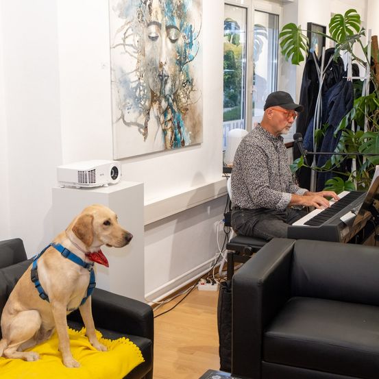 Ein Mann spielt an einem Tisch eine Tastatur, während ein Hund in einem Geschirr auf einem Stuhl sitzt. Ein Projektor und ein Gemälde stehen auf einem Ständer.