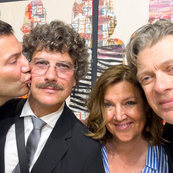 Drei Personen posieren für ein Selfie vor einem farbenfrohen, abstrakten Kunstwerk. Der Mann auf der linken Seite küsst den Mann in der Mitte auf die Wange. Die Frau auf der rechten Seite lächelt.