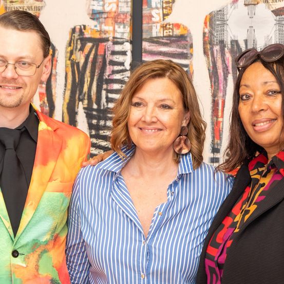 Drei Menschen stehen vor abstrakter Kunst. Ein Mann mit Brille, eine Frau mit gestreifter Bluse und eine weitere Frau mit einem gemusterten Mantel. Sie alle lächeln für die Kamera.