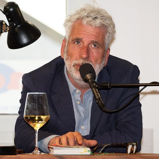 Ein älterer Mann mit weißem Haar und Bart sitzt an einem Tisch mit einem Mikrofon und einem Weinglas, er trägt einen dunkelblauen Anzug.