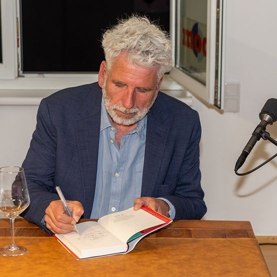 Ein Mann sitzt an einem Tisch und unterschreibt ein Buch mit einem Stift. Ein Glas Wein und eine Lampe sind auf dem Tisch. Ein Mikrofon ist hinter ihm positioniert.