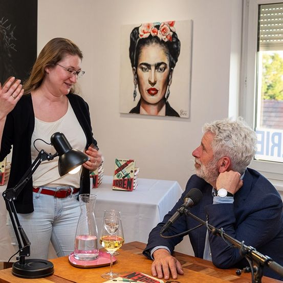 Ein Mann und eine Frau sitzen mit Mikrofonen an einem Tisch. Sie haben eine Lampe, Bücher und Weingläser. Hinter ihnen befindet sich ein Gemälde von Frida Kahlo und ein Porträt von Jesus.