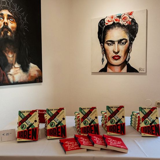 Ein Tisch mit mehreren Büchern mit dem Titel Irren, eines zum Preis von 25 Euro, umgeben von Kunst. Ein Gemälde zeigt Jesus und das andere Frida Kahlo.