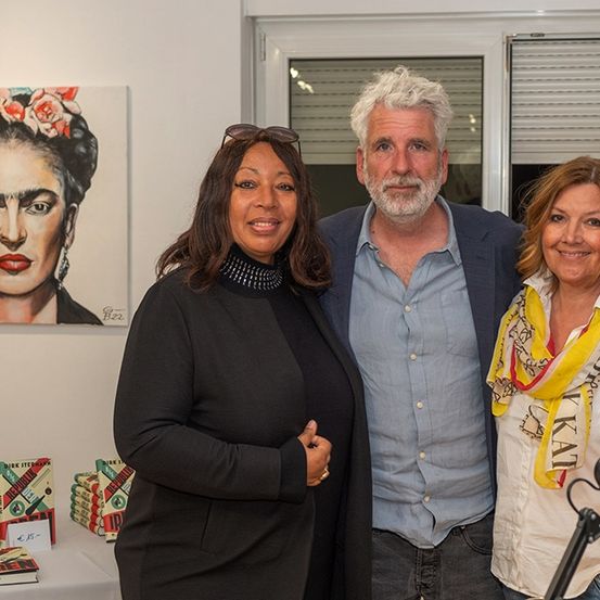 Drei Personen stehen bei einer Kunstausstellung zusammen. Der Mann hat weiße Haare und einen Bart, während die beiden Frauen Schals tragen. Hinter ihnen befindet sich ein Bild von Frida Kahlo.