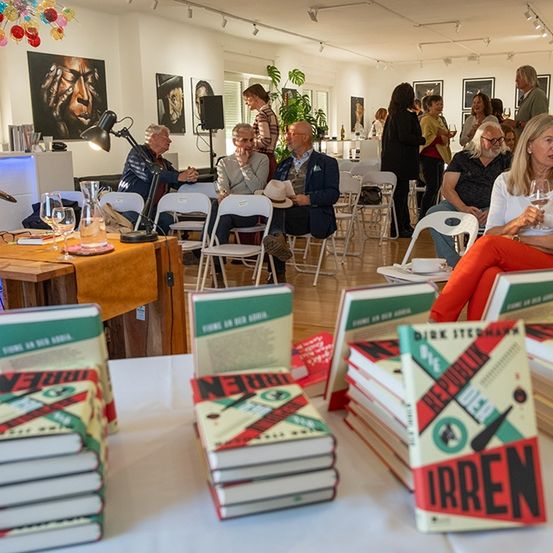 Ein Raum voller Menschen, die in Stühlen sitzen, einige lesen Bücher, wahrscheinlich bei einer Veranstaltung. Ein Tisch im Vordergrund hat mehrere Bücher, die darauf gestapelt sind, eines mit dem Titel Irgen.