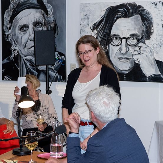 Eine Frau steht zwischen zwei großen Gemälden und spricht zu einem sitzenden Publikum. Ein Mann hört aufmerksam zu, während eine andere Frau ein Glas Wein hält.