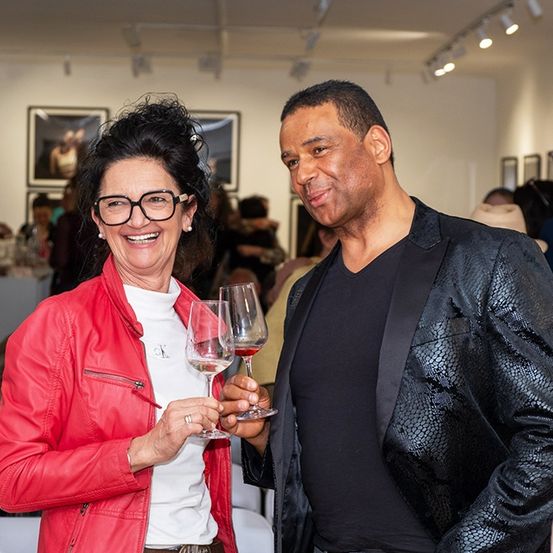 Ein Mann und eine Frau halten Weinglas, lächeln, bei einer Veranstaltung in einer Galerie. Andere Leute, Kunstwerke und ein Tisch mit Gegenständen sind im Hintergrund.