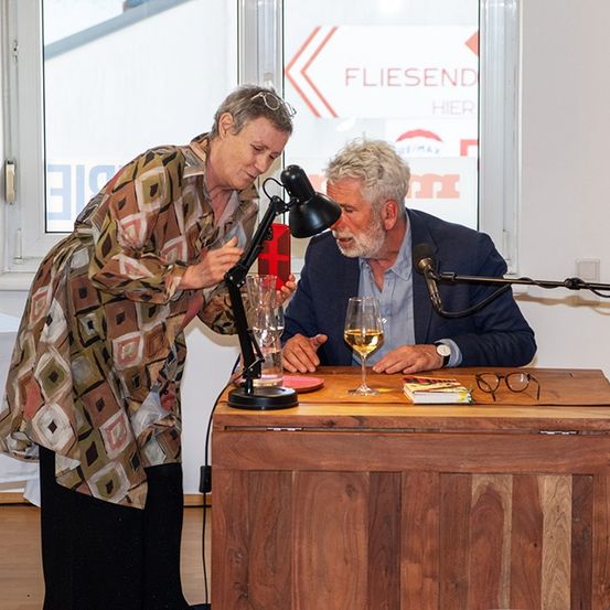 Ein Mann und eine Frau stehen hinter einem Holzpodium. Die Frau zeigt auf eine Lampe und der Mann schaut hin, beide mit Weingläsern auf dem Tisch.