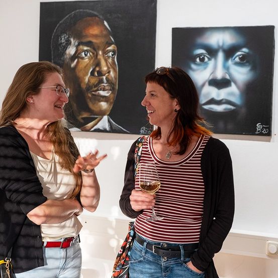 Zwei Frauen unterhalten sich vor zwei großen Porträts an einer weißen Wand. Eine hält ein Weinglas.