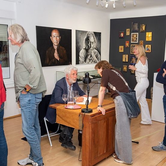 Eine Gruppe von Menschen ist um einen Tisch mit einem sitzenden Mann versammelt, möglicherweise ein Künstler. Sie befinden sich in einem Raum mit Kunstwerken an der Wand und einem Tisch mit Weinläsern und einem Buch.