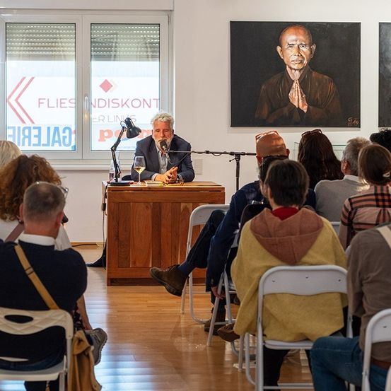 Ein Mann spricht an einem Podium in einer Kunstgalerie, umgeben von Stühlen, mit einem Publikum und großen Gemälden im Hintergrund.