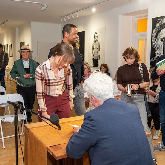 Ein Mann signiert ein Buch an einem Podium mit Mikrofon, während mehrere Leute stehen und zuhören. Eine Kunstgalerie ist im Hintergrund.