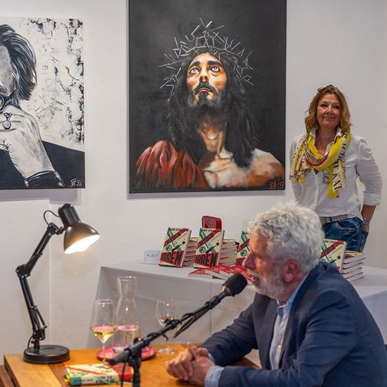 Ein Mann sitzt mit einem Mikrofon an einem Tisch und schaut eine Frau an. Ein Gemälde von Jesus hängt an der Wand.