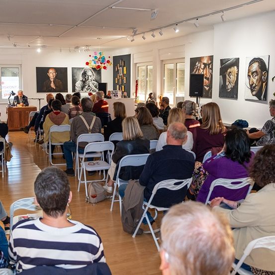 Eine Kunstgalerie beherbergt ein Publikum, das in Stühlen sitzt und einem Mann zuhört, der auf der Bühne spricht. Porträts schmücken die Wände, und Deckenbeleuchtung erhellt den Raum.
