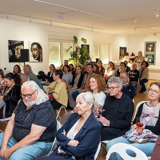 Eine Gruppe von Menschen sitzt in einem Raum mit Kunstwerken an der Wand, einige mit Brille, Weinglas in der Hand und schaut etwas an. Der Raum hat weiße Wände, eine weiße Decke und ein Fenster.