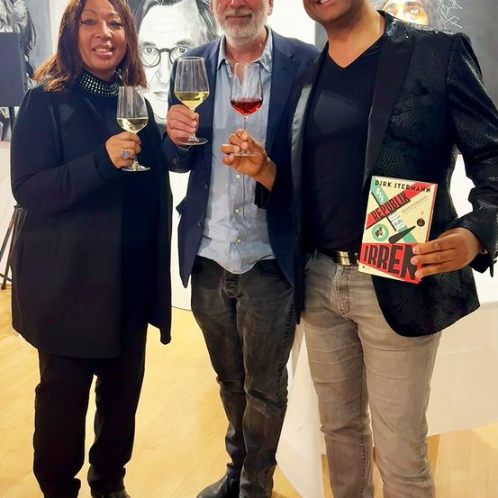 Drei Personen lächeln in einem Innenbereich, möglicherweise in einer Galerie. Die Frau auf der linken Seite hält ein Weinglas, und der Mann neben ihr hält zwei. Der Mann auf der rechten Seite hält ein Buch. Hinter ihnen befindet sich ein Gemälde an der Wand.