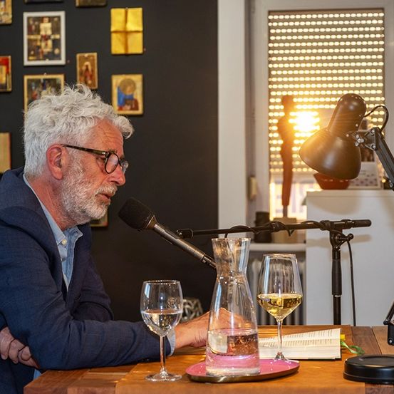 Ein älterer Mann im Anzug sitzt an einem Tisch mit Weingläsern und einer Lampe. Er spricht in ein Mikrofon.