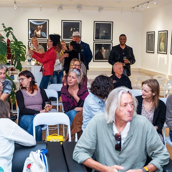 Eine Gruppe von Menschen ist in einem Raum, einige sitzen und einige stehen, alle schauen auf eine Wand mit mehreren Kunstrahmen, die daran hängen. Einige von ihnen halten Weingläser.