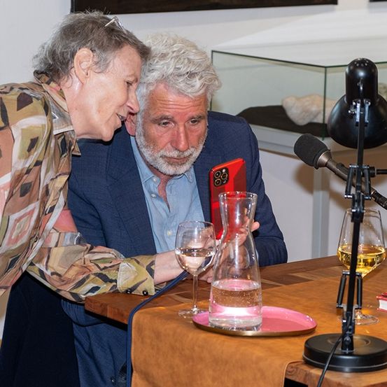 Ein älterer Mann und eine ältere Frau sitzen zusammen, er hält ein rotes Telefon, beide sehen amüsiert aus. Gläser, eine Weinkaraffe und ein Buch sind auf dem Tisch, mit einem Mikrofon in der Nähe.