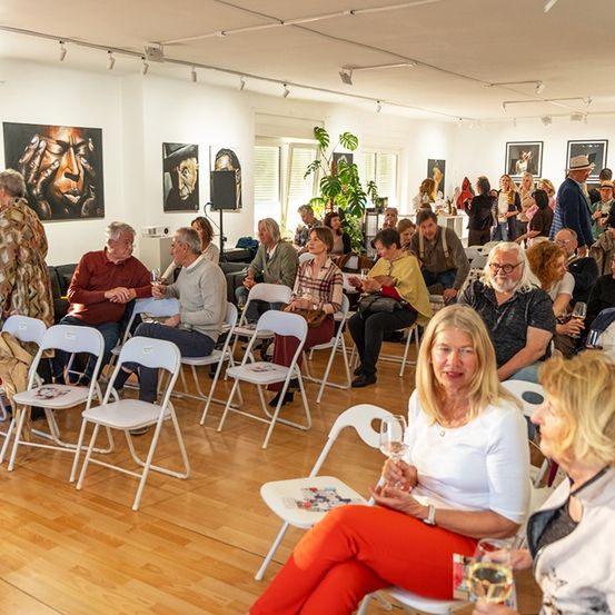 Ein Raum voller Menschen, die auf Stühlen sitzen, wobei einige in der Nähe einer Wand mit Kunstwerken stehen. Der Raum ist gut beleuchtet mit Deckenlampen. Einige Personen führen Gespräche und einige halten Weingläser in der Hand. Kunstwerke schmücken die Wände.