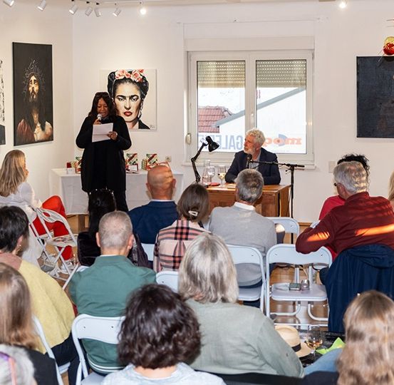 Eine Frau steht mit einem Mikrofon an einem Podium in einem Raum voller Menschen, die auf Stühlen sitzen, ein Mann sitzt an einem Schreibtisch mit einem Glas, Gemälde hängen an den Wänden und Lampen sind an der Decke angebracht.