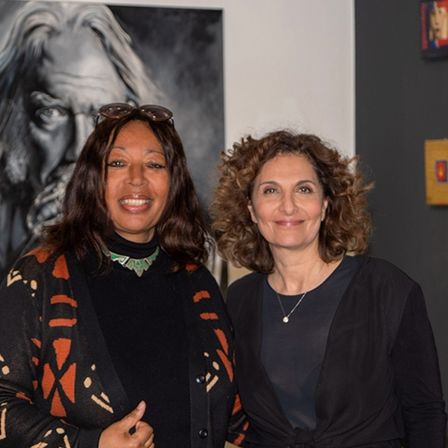 Zwei Frauen stehen Seite an Seite vor einer Wand mit gerahmten Kunstwerken. Eine Frau lächelt mit der Hand auf der Brust, und die andere trägt eine Sonnenbrille auf dem Kopf. Dahinter befindet sich ein großes schwarz-weißes Gemälde eines bärtigen Mannes.