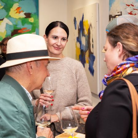 Eine Frau in einem grauen Pullover und Hut unterhält sich mit zwei anderen, die Weingläser halten. Dahinter hängen abstrakte Gemälde an der Wand.