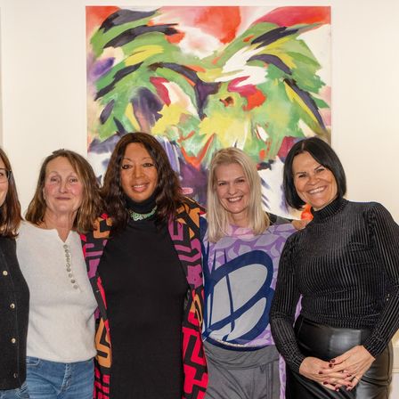 Fünf Frauen stehen zusammen vor einem abstrakten Gemälde. Die Frau ganz rechts trägt ein schwarzes Rollkragenshirt und Lederhosen.
