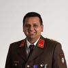 Ein lächelnder Mann in Uniform mit mehreren Medaillen auf der Brust, vor weißem Hintergrund.