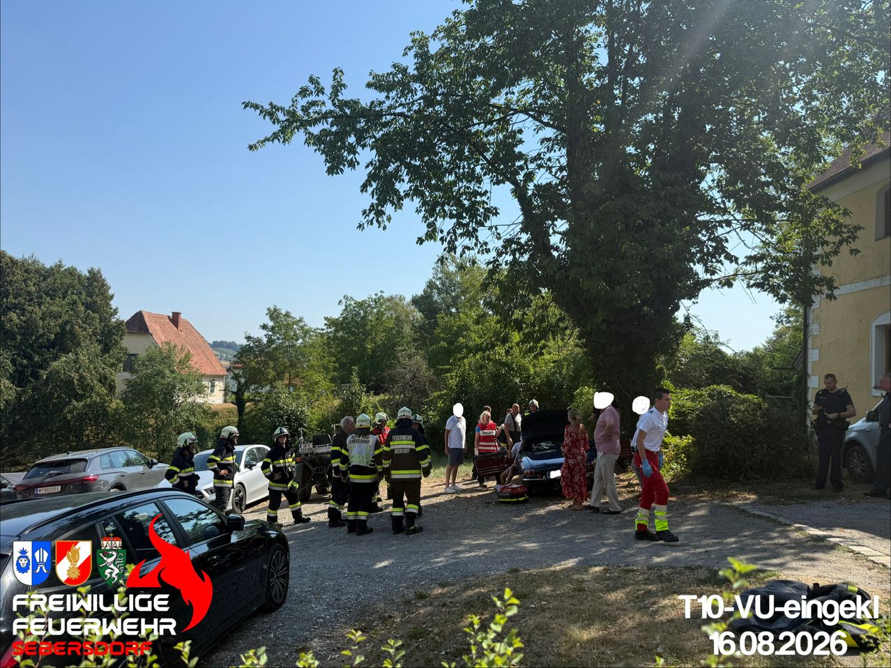 Einsatzkräfte, darunter Feuerwehrleute, versammeln sich um ein beschädigtes Fahrzeug auf einer Schotterstraße. In der Nähe beobachten eine Gruppe von Menschen die Szene. Bäume und ein Haus sind im Hintergrund zu sehen.