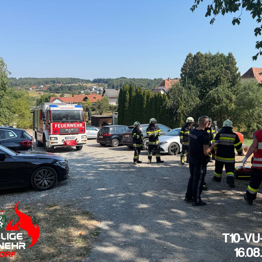 Eine Gruppe von Feuerwehrleuten und Polizeibeamten steht auf einem Parkplatz mit einem Feuerwehrwagen und Autos. Die Feuerwehrleute tragen Helme und Uniformen. Der Polizeibeamte trägt ein schwarzes Hemd und eine Hose. Der Feuerwehrwagen steht in der Mitte des Parkplatzes.