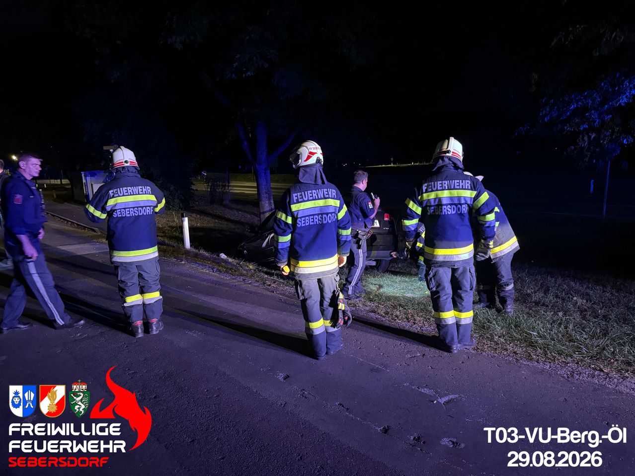 Vier Feuerwehrleute in blauen Uniformen mit gelben Streifen stehen bei Nacht am Straßenrand. Einer von ihnen schaut auf ein Auto, das am Straßenrand geparkt ist.