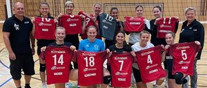 Eine Volleyballmannschaft von Frauen und einem Trainer posiert für ein Foto auf einem Platz und hält rote Trikots mit Nummern. Sie stehen vor einem Netz und einer Wand mit Fenstern.